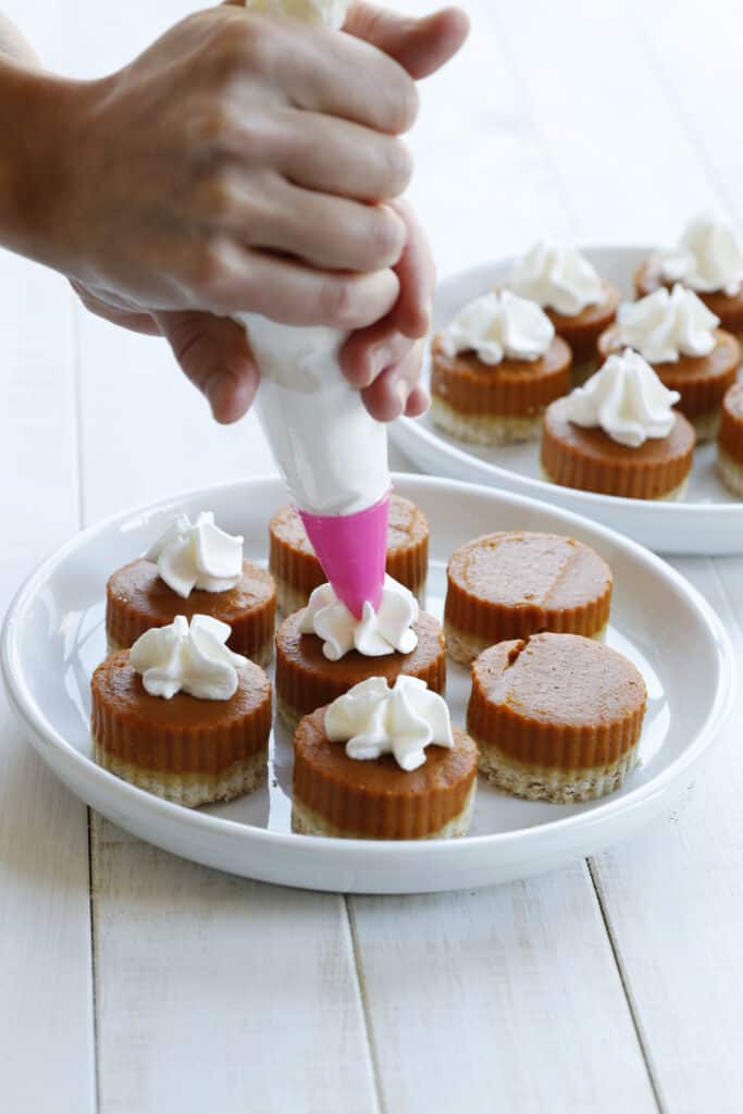 Someone putting whipping topping from a piping bag onto vegan pumpkin pie bites on a plate.