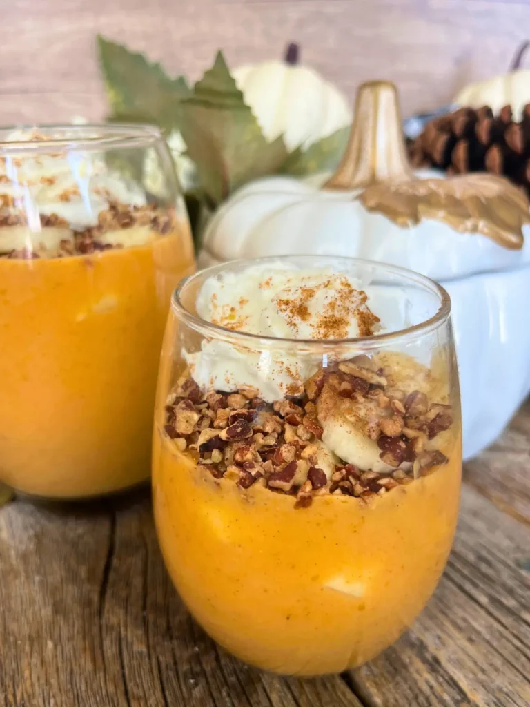 Two healthy pumpkin cheesecake puddings in glass jars on a counter next to a glass pumpkin.