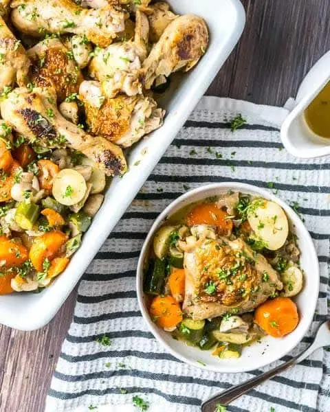 A bowl of chicken and vegetables next to a sheet pan of the chicken and vegetable dinner on a table.