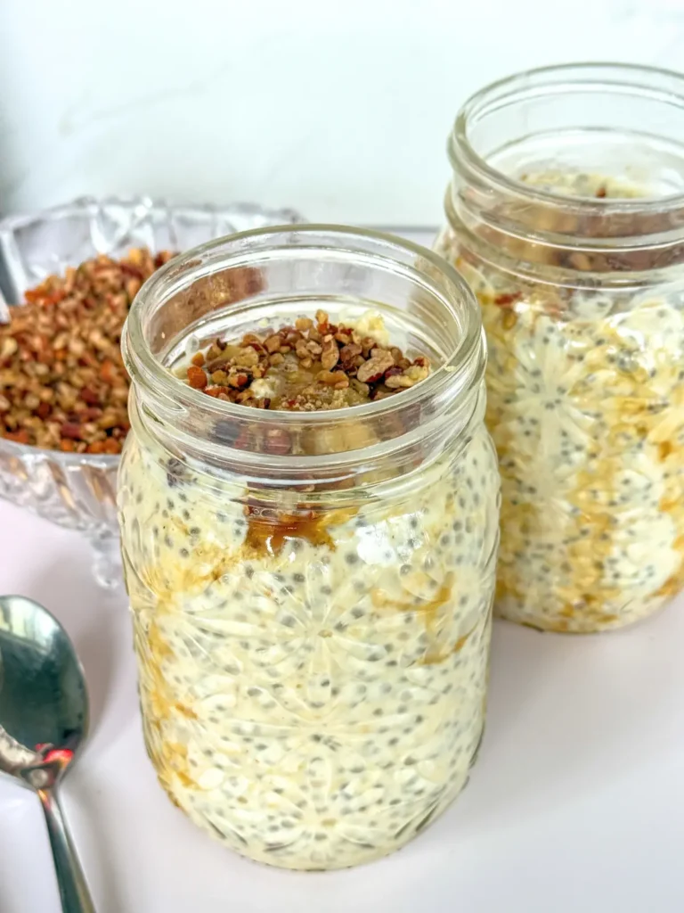 Two open mason jars of Greek yogurt overnight oats topped with maple, brown sugar, and chopped pecans next to a spoon and a bowl of pecans.