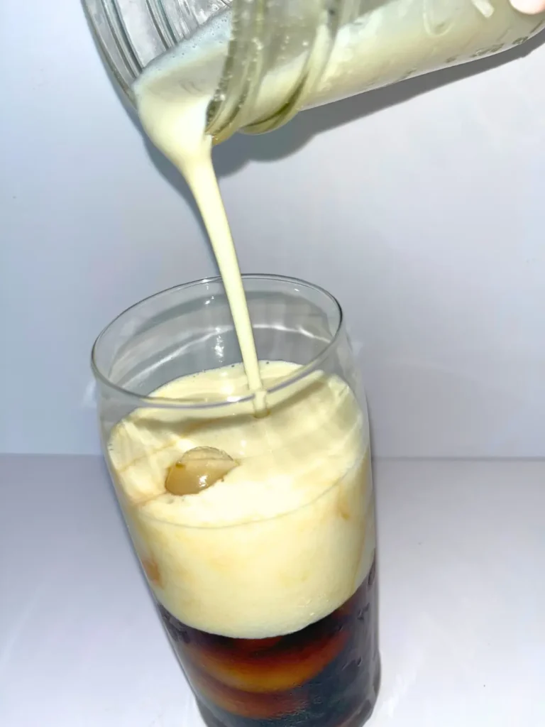 Pouring the thick protein cold foam into a glass of iced coffee.