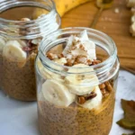 Two glass jars filled with pumpkin cheesecake protein pudding topped with banana slices and chopped pecans.