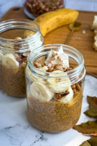 Two glass jars filled with pumpkin cheesecake protein pudding topped with banana slices and chopped pecans.