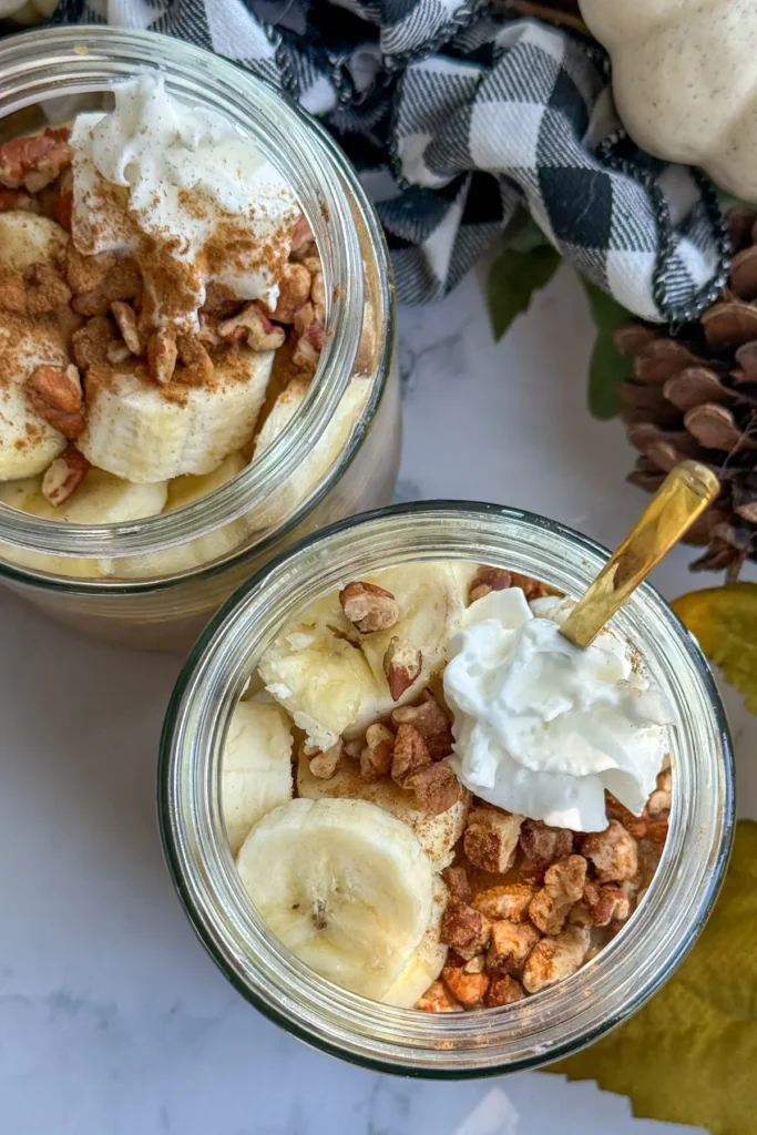 A birds-eye view of two jars of protein pudding topped with whipped cream, banana slices, and chopped pecans on a counter decorated with fall decor.