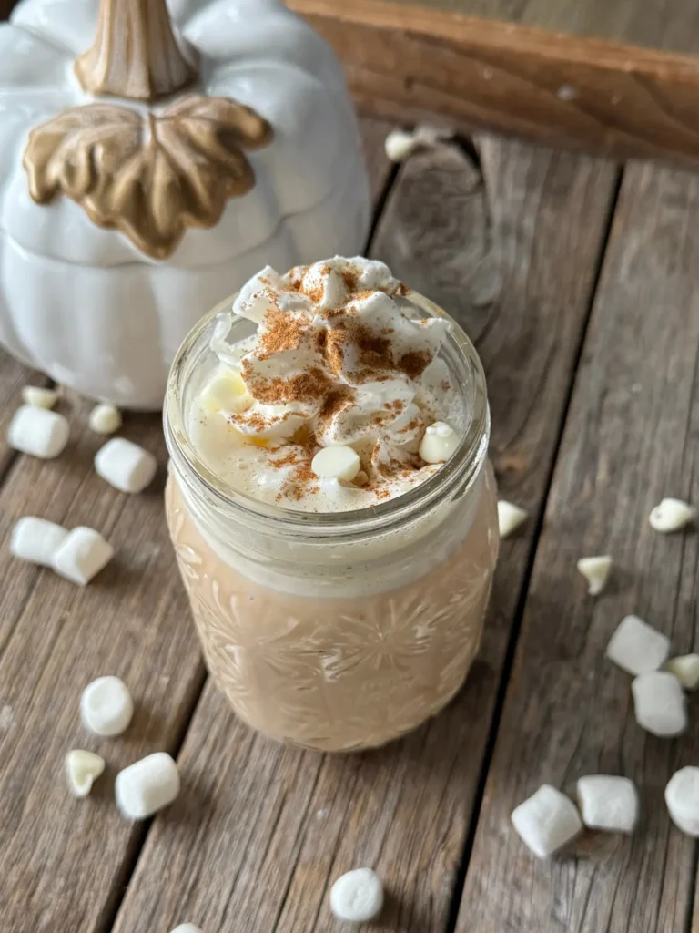 Pumpkin spice hot cocoa in a glass mug, topped with whipped cream and pumpkin pie spice on a wooden cutting board.