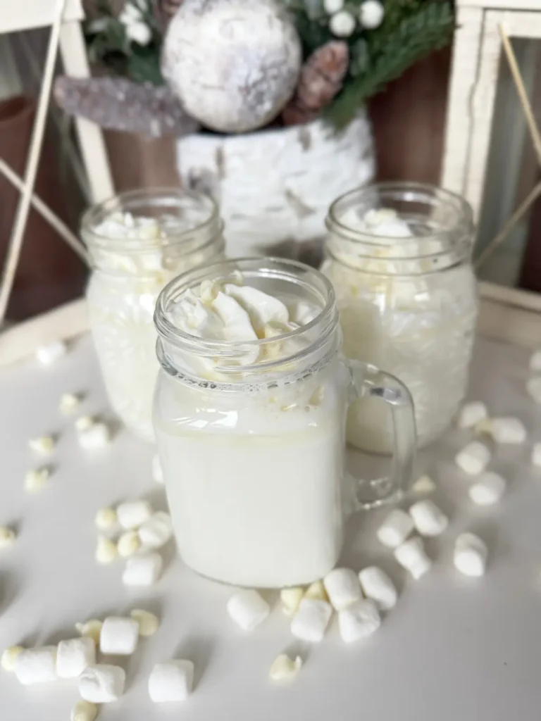 Three glass mugs filled with white chocolate and marshmallow cream hot cocoa with decorative lanterns and a winter themed pot in the background.