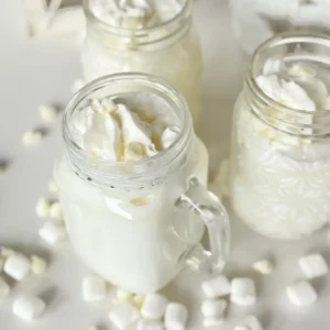 Three white chocolate hot chocolates with marshmallow cream in glass mugs on a counter decorated with mini marshmallows and white chocolate chips.