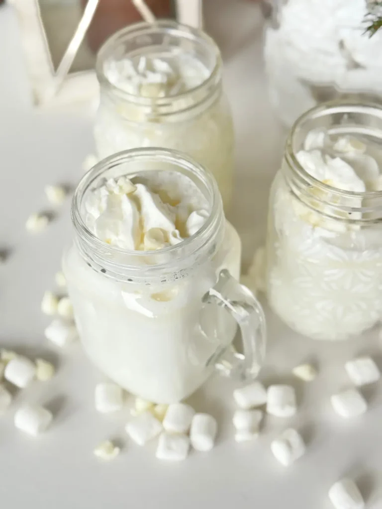 Three white chocolate hot chocolates with marshmallow cream in glass mugs on a counter decorated with mini marshmallows and white chocolate chips.