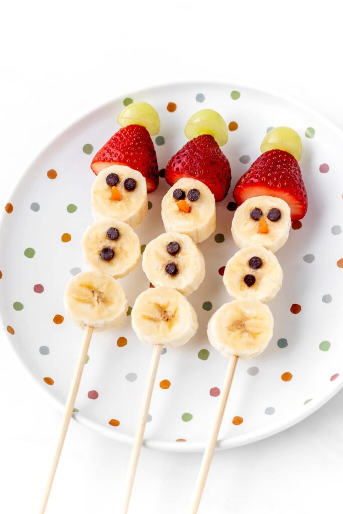 Three banana snowmen fruit kabobs on a polka dotted plate.