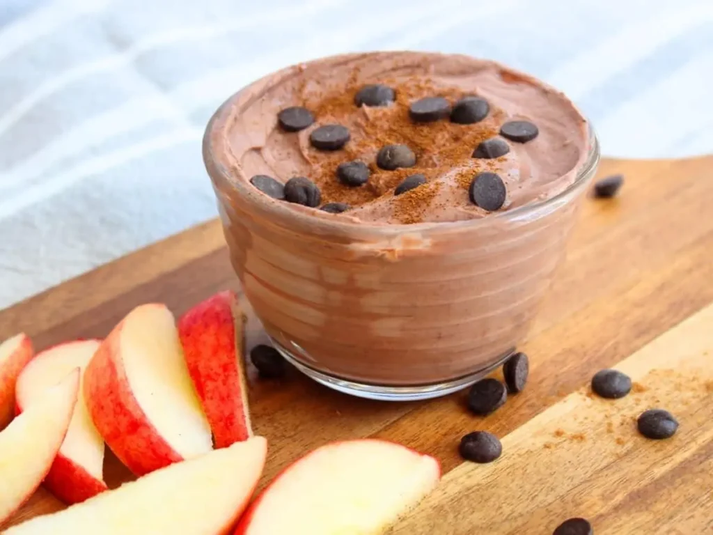 Brownie batter dip topped with chocolate chips on a cutting board next to apple slices.