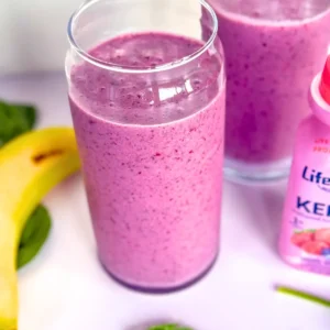 Two banana berry smoothies in glasses on a counter next to a banana and a bottle of kefir.