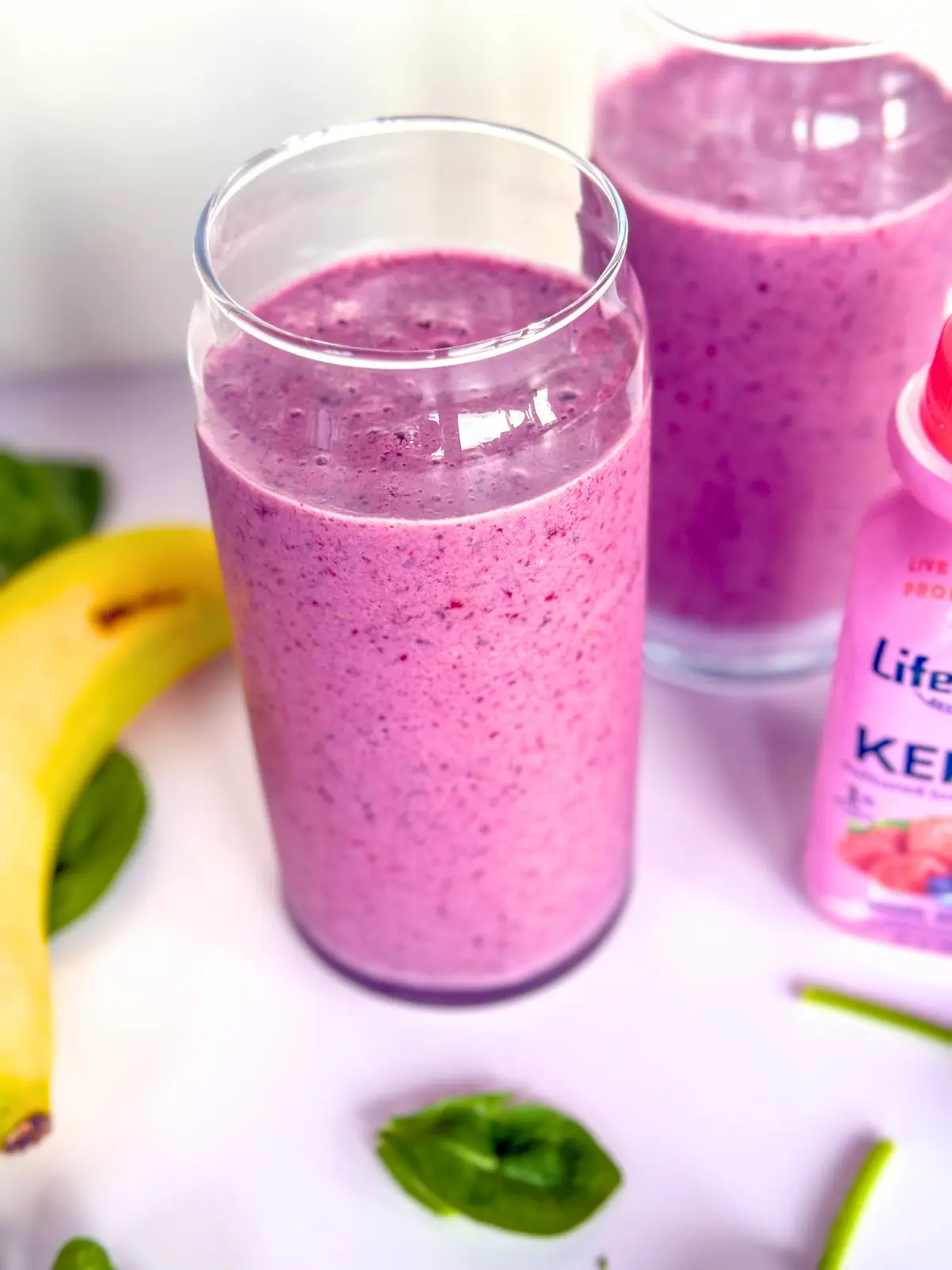 Two banana berry smoothies in glasses on a counter next to a banana and a bottle of kefir.