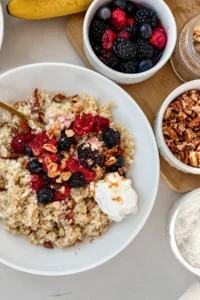 A creamy coconut quinoa bowl topped with mixed berries, chopped pecans, and whipped cream on a table next to berries, bananas, and pecans.