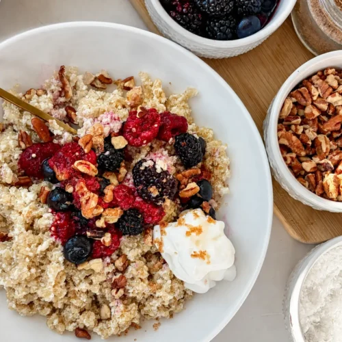 A creamy coconut quinoa bowl topped with mixed berries, chopped pecans, and whipped cream on a table next to berries, bananas, and pecans.