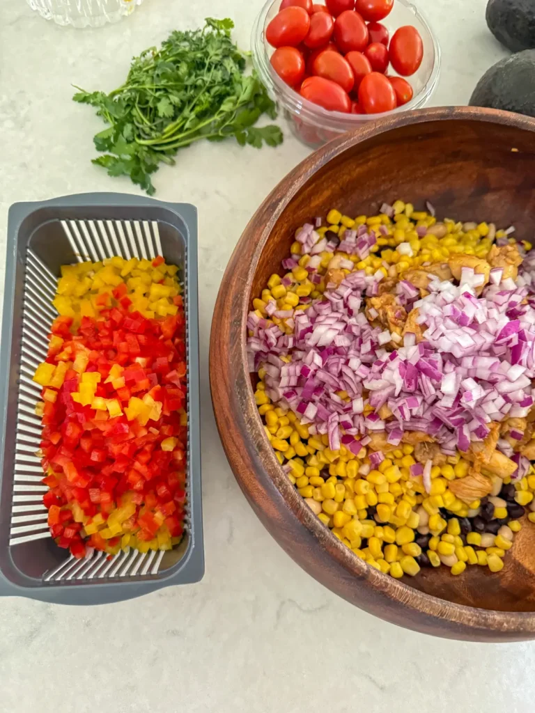 A vegetable chopper with diced bell peppers in it next to a large bowl of onions, beans and corn.