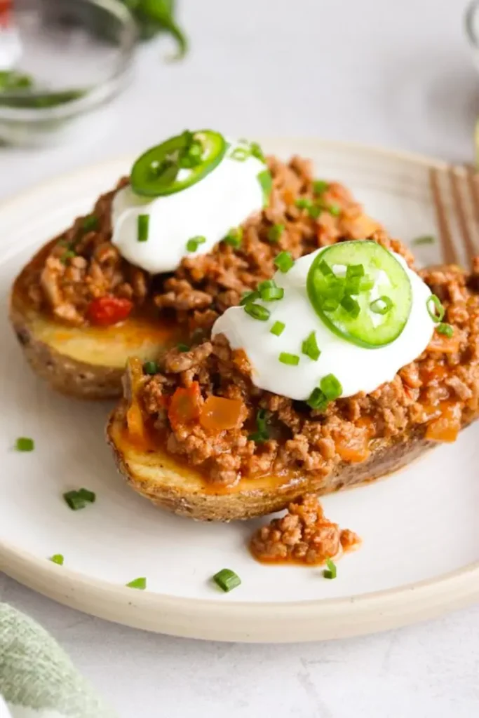 Two baked potatoes topped with taco meat, sour cream and jalapenos on a plate next to a fork on the table.