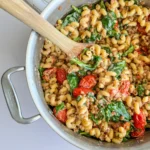 A large pot of high protein feta, tomato and lentil pasta with a wooden spoon in it on a decorative towel.