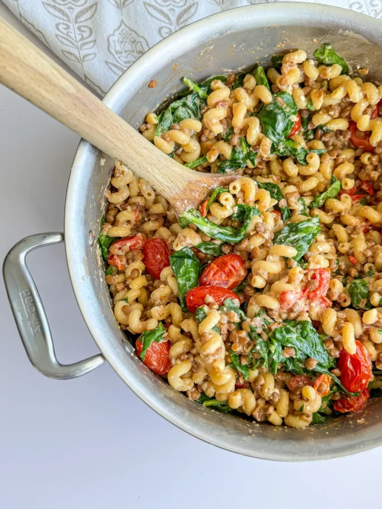 A large pot of high protein feta, tomato and lentil pasta with a wooden spoon in it on a decorative towel.