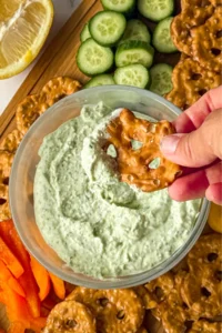 A pretzel chip with bsail parmesan yogurt dip on it help over a bowl of dip on a snack board.