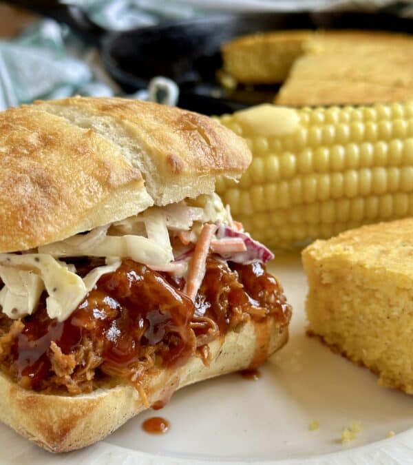 A slow cooker pulled pork sandwich on a plate next to corn and cornbread.