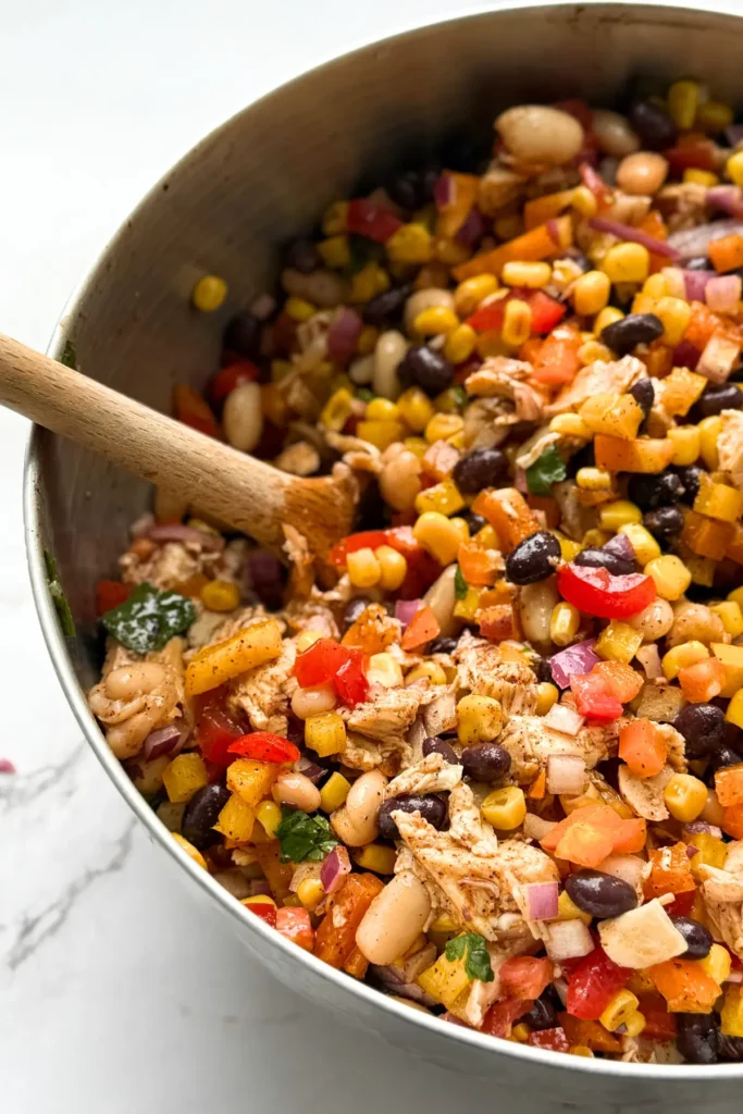 A large stainless steel bowl of Mexican dense bean salad on a counter with a wooden spoon in it.