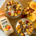 A birds-eye view of two bowls of fully assembled peach cobbler overnight oats on a wooden cutting board next to a bottle of kefir and some diced peaches.