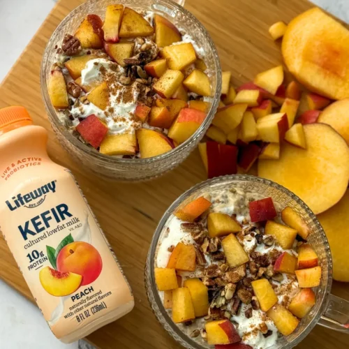 A birds-eye view of two bowls of fully assembled peach cobbler overnight oats on a wooden cutting board next to a bottle of kefir and some diced peaches.