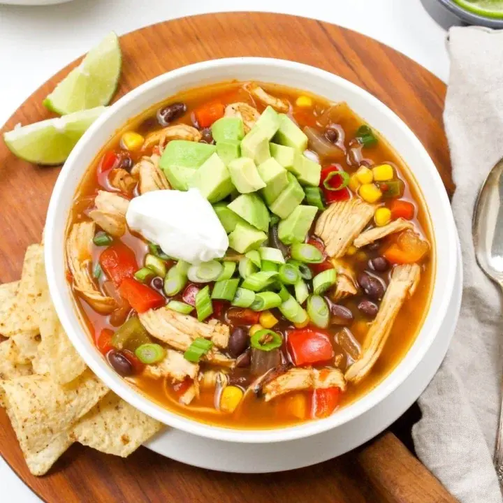 A bowl of slow cooker chicken taco soup on a wood platter next to chips and lime wedges.