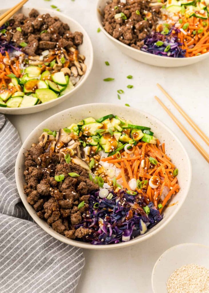 Fully assemble Korean ground beef bowls with raw vegetables and rice on a table next to chopsticks.