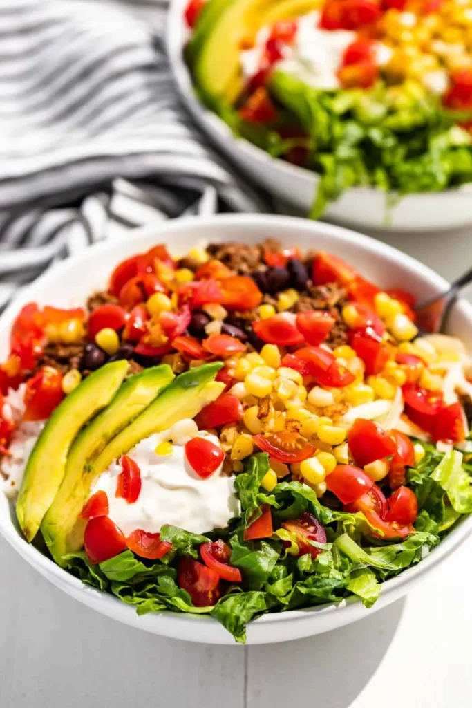 A ground beef taco bowl with avocado slices, tomatoes, corn and shredded lettuce in a bowl on a table.