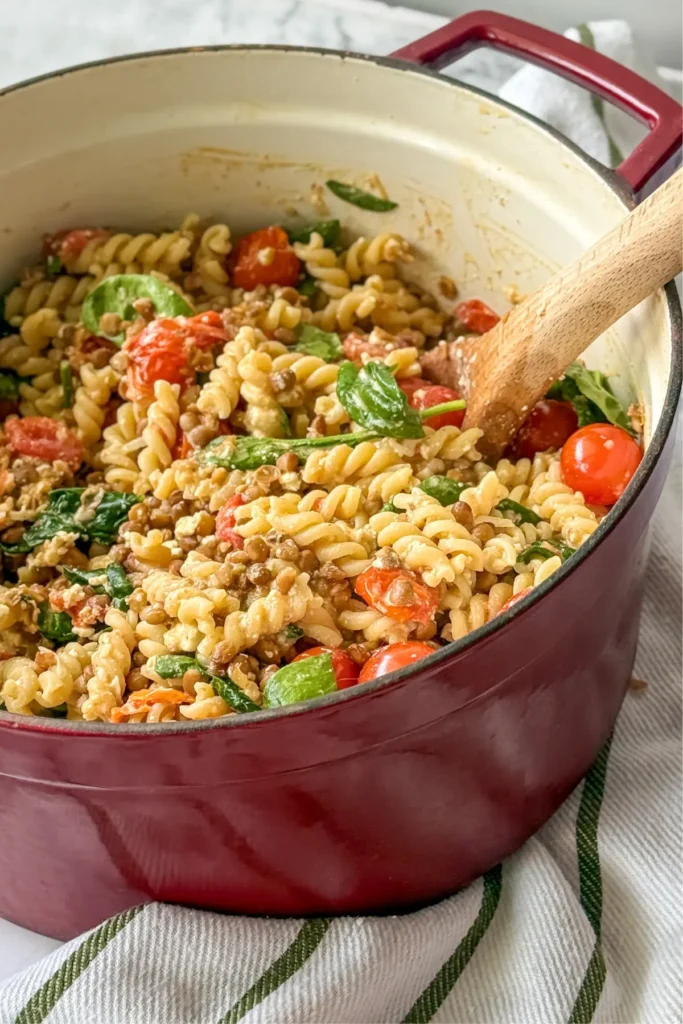 A big pot of feta and tomato pasta with lentils and spinach with a wooden spoon in it on a counter next to a towel.