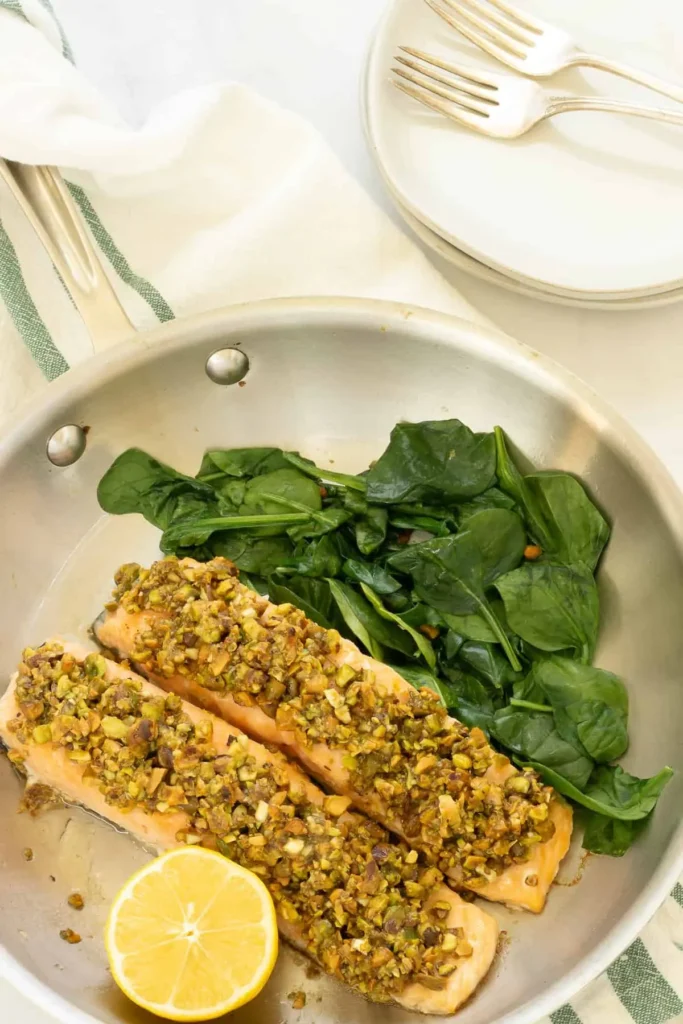 Pistachio crusted salmon filets in a pan next to spinach and a lemon wedge.