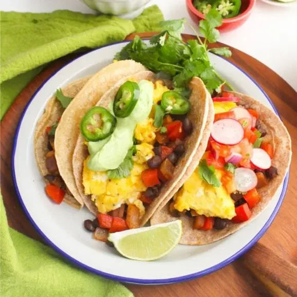 Three veggie and egg breakfast tacos with jalapenos on a plate on a wooden table.