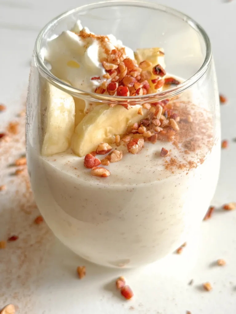 A small glass of cinnamon roll pudding with protein powder topped with banana slices, whipped cream, and chopped pecans.