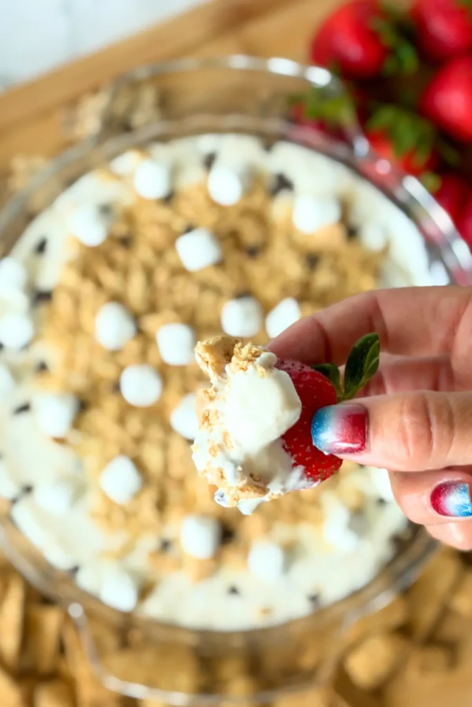 A close-up of a strawberry dipped into the s'mores dip over a circular round glass dish on a dessert board.