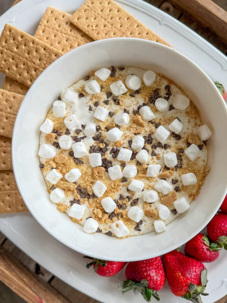 A large bowl of s'mores dip with cottage cheese on a platter surrounded by graham crackers and strawberries.