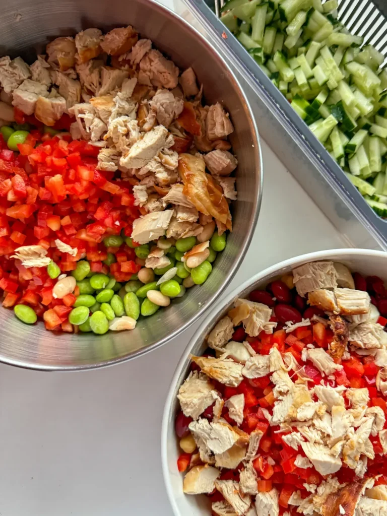 Two bowls with chopped veggies, chicken, and beans on a counter next to a vegetable chopper with diced cucumber in it.