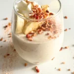 A small glass of cinnamon roll pudding with protein powder topped with banana slices, whipped cream, and chopped pecans on a counter.