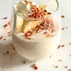 A small glass of cinnamon roll pudding with protein powder topped with banana slices, whipped cream, and chopped pecans on a counter.