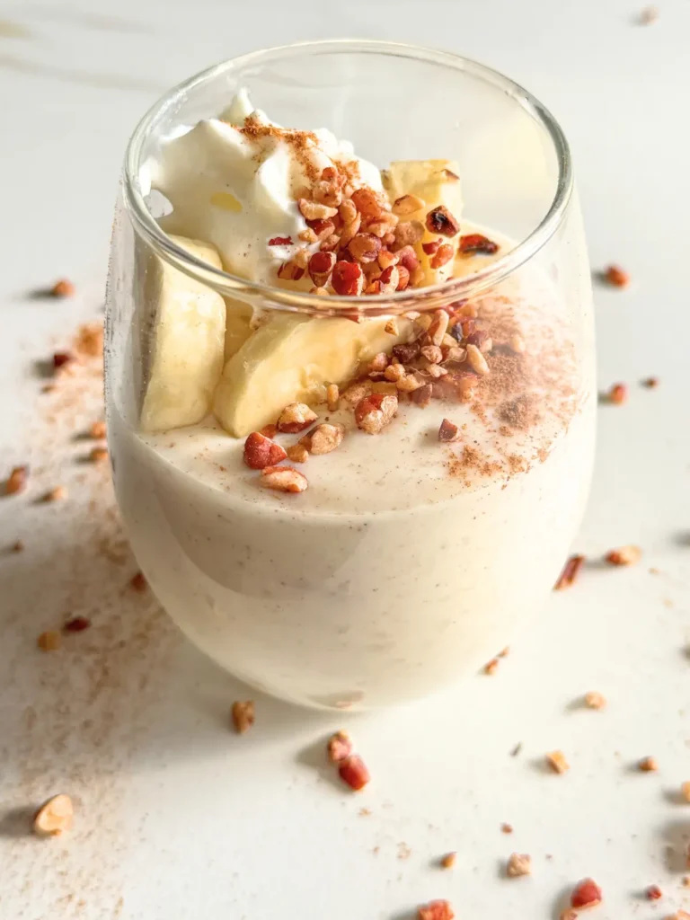 A small glass of cinnamon roll pudding with protein powder topped with banana slices, whipped cream, and chopped pecans on a counter.