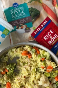 A large pan of healthier Rice-A-Roni on a counter next to an empty box of Rice-A-Roni and frozen vegetables.