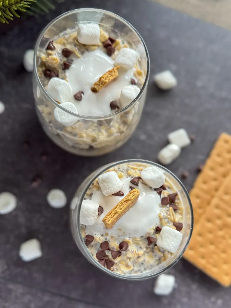 A birds-eye view of two glasses of s'mores overnight oats topped with marshmallow cream and graham cracker crumbs on a counter surrounded by crushed graham cracker and mini marshmallow.