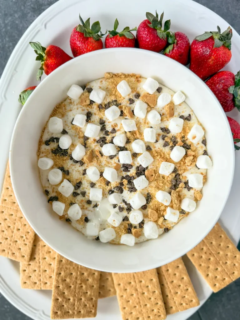 A fully assembled cottage cheese s'mores dip in a bowl on a white platter with graham cracker sheets and strawberries.