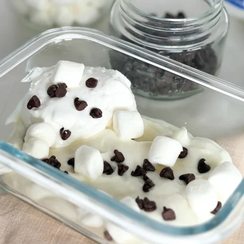 A snack container with s'mores Greek yogurt topped with marshmallow cream and chocolate chips on a counter.