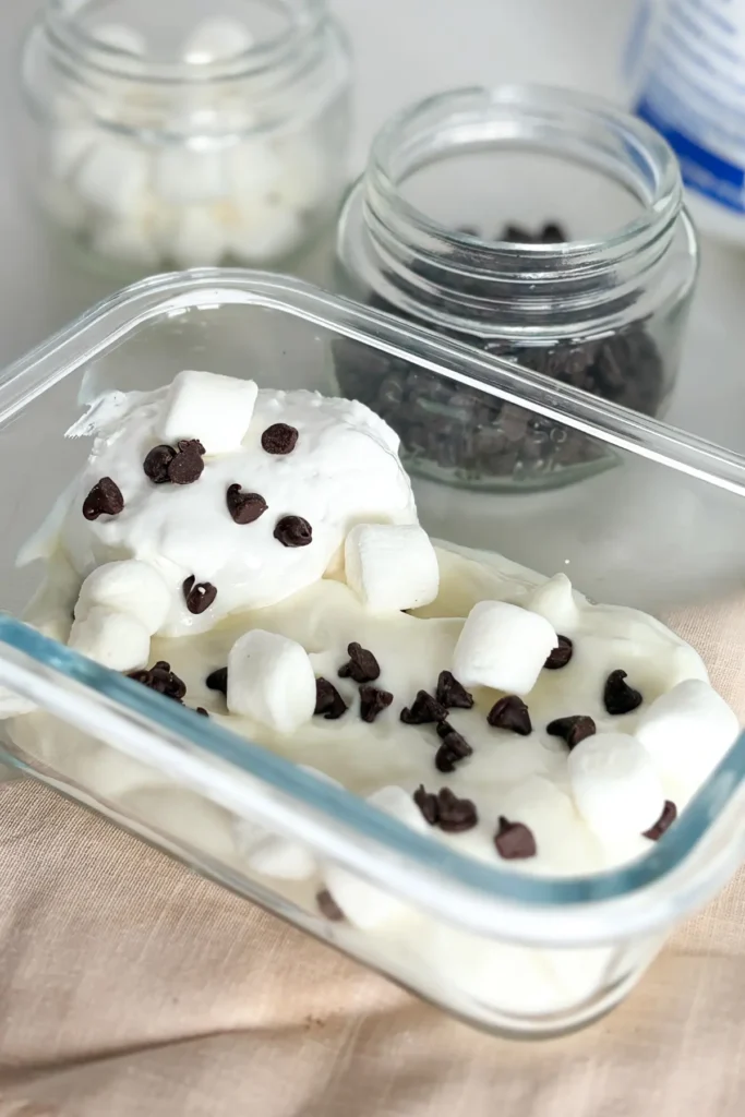 A snack container with s'mores Greek yogurt topped with marshmallow cream and chocolate chips on a counter.