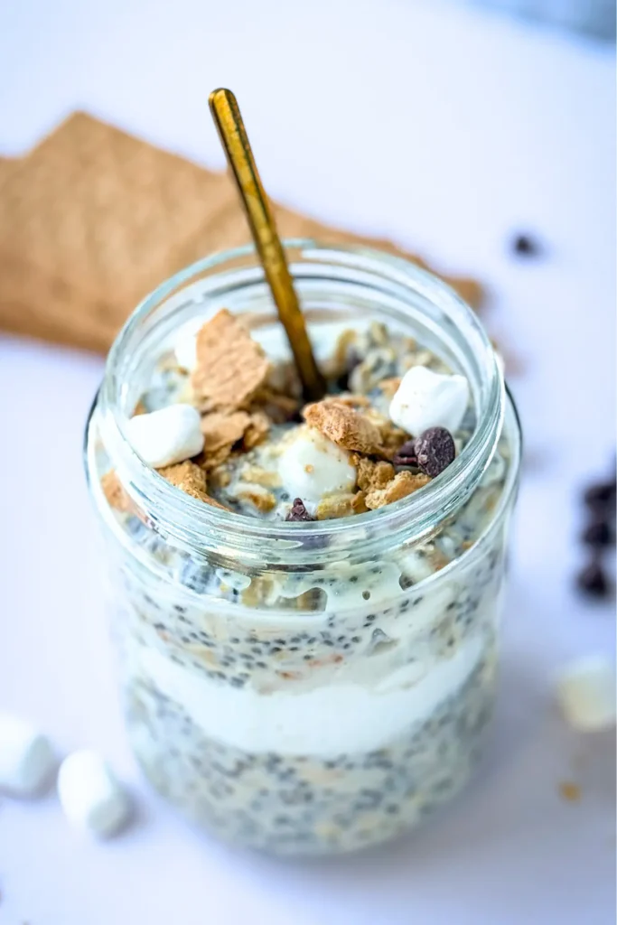 A fully assembled s'mores overnight oats in a jar with a gold spoon in it on a counter next to a graham cracker and mini marshmallows.