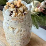 A large mason jar with s'mores overnight oats topped with crushed graham crackers, mini marshmallows, marshmallow cream and chocolate chips on a small wooden cutting board.