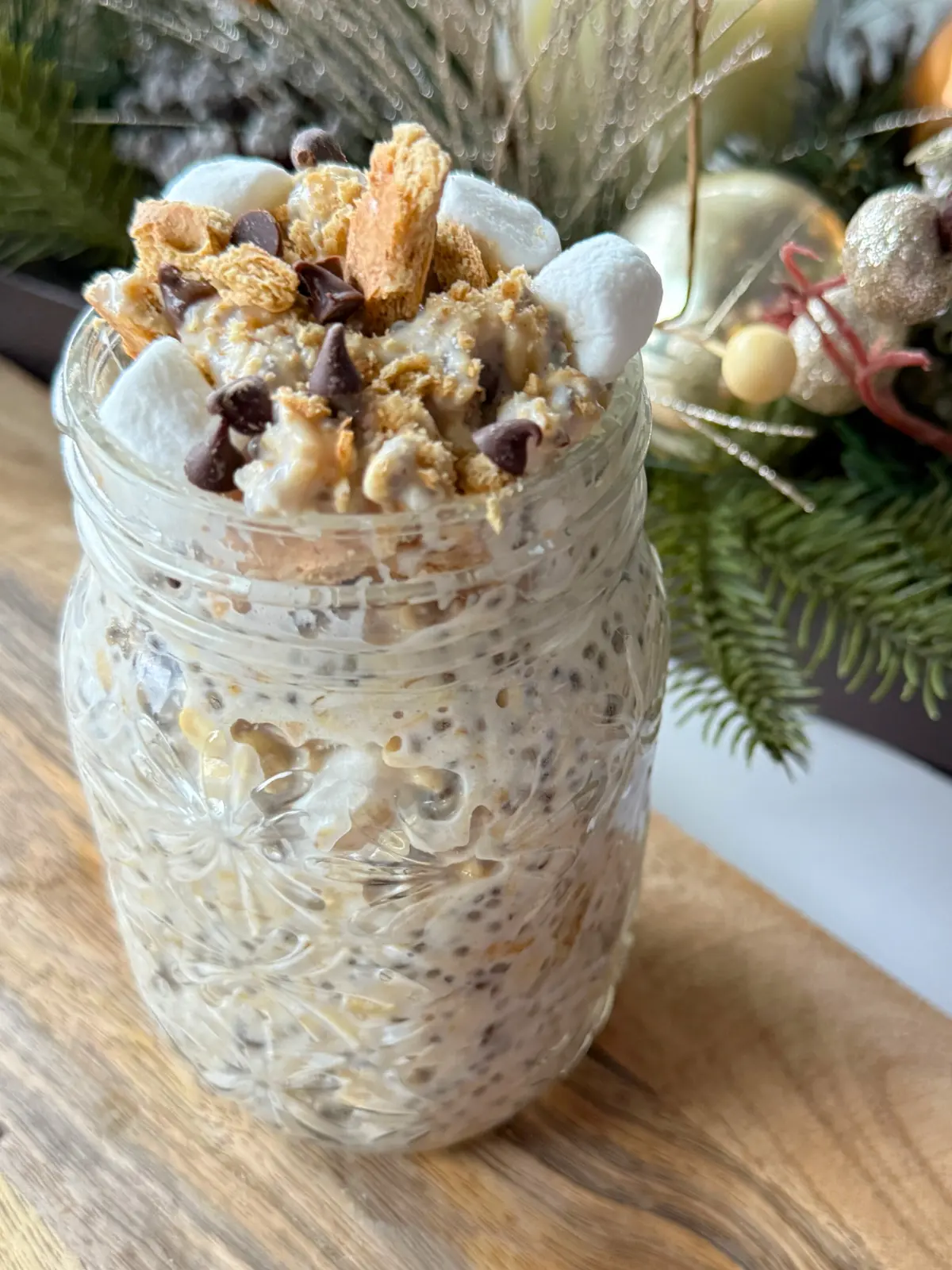 A large mason jar with s'mores overnight oats topped with crushed graham crackers, mini marshmallows, marshmallow cream and chocolate chips on a small wooden cutting board.