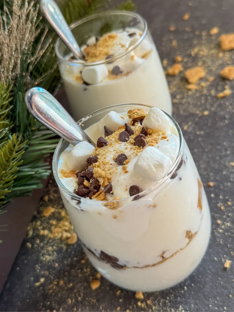 Two glasses full of s'more protein pudding made with Greek yogurt topped with marshmallow cream, crushed graham crackers and mini chocolate chips on a counter.