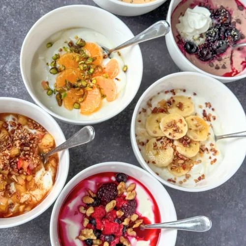 6 fully assembled yogurt bowls on a counter with spoons in the bowl.